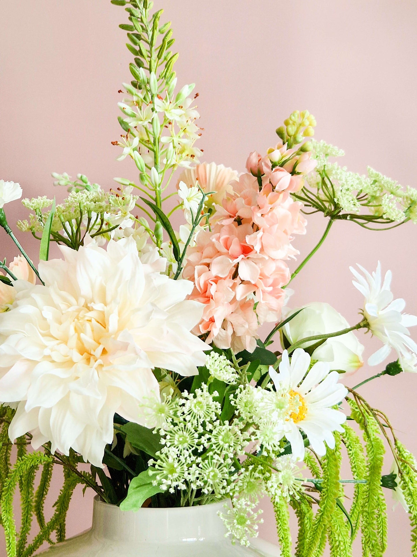Close-up van een zijden bloemenboeket met witte dahlia's, roze Matthiola, madeliefjes en schermbloemen in een witte vaas tegen een roze achtergrond.