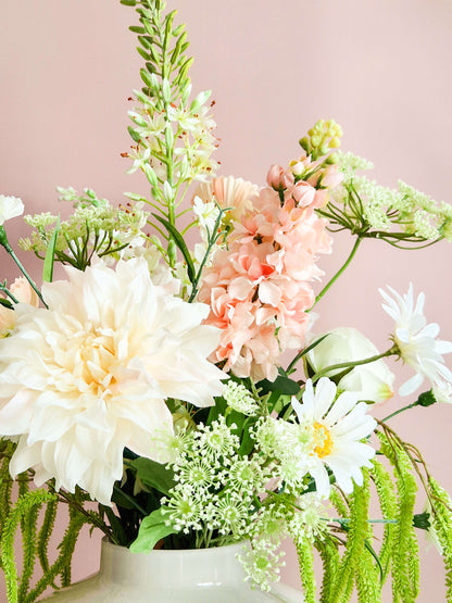 Close-up van een zijden bloemenboeket met witte dahlia's, roze Matthiola, madeliefjes en schermbloemen in een witte vaas tegen een roze achtergrond.
