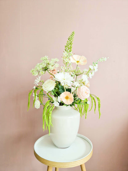 Stijlvol zijden bloemenboeket in zachte tinten wit, roze en groen in een elegante vaas op een rond houten tafeltje tegen een lichtroze muur.