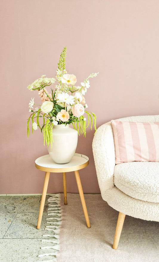 Zijden bloemenboeket met pastelkleuren op een tafeltje naast een crèmekleurige bank met roze kussen in een sfeervol interieur.